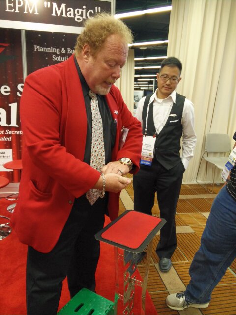 trade-show-magician-candid-booth-interaction Trade show magician in a red blazer talking with a booth attendee at a convention exhibit