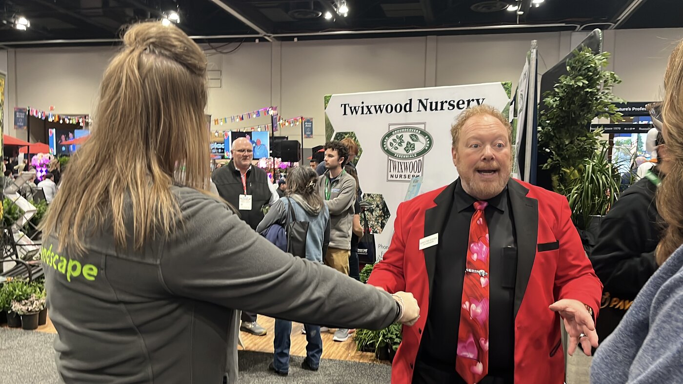 trade-show-magician-engaging-attendee-mid-conversation Trade show magician in a red blazer talking with an attendee on the show floor at a landscape and nursery trade show