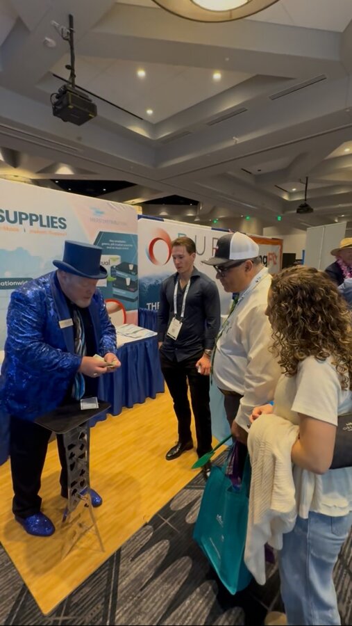 trade-show-magician-engaging-booth-visitors Trade show magician performing a close-up card trick for attendees gathered at a convention booth